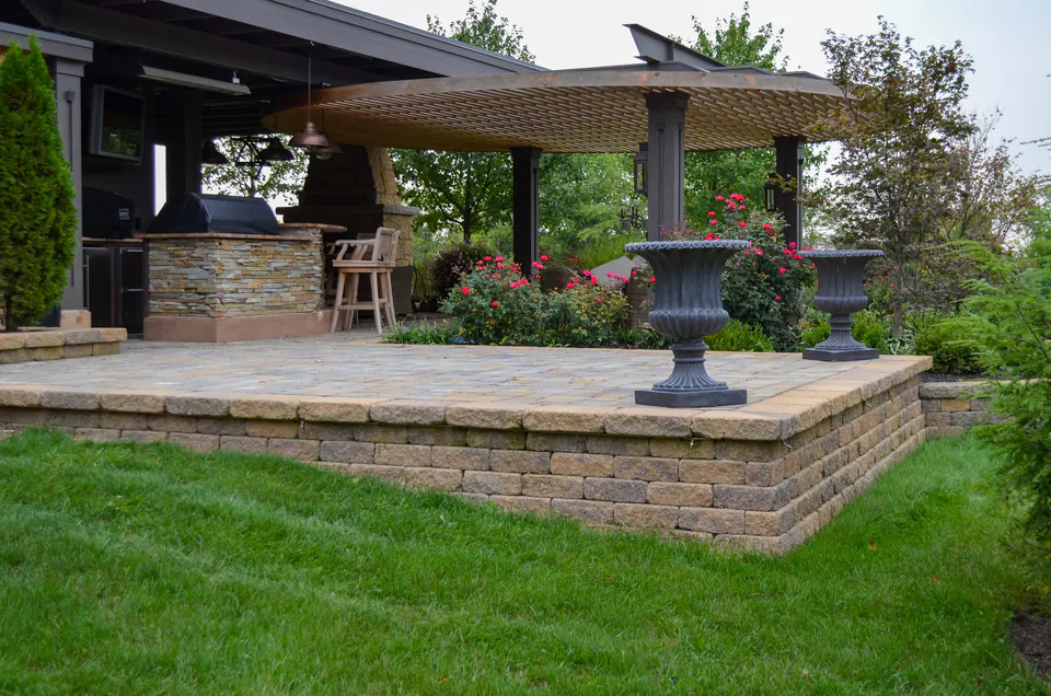 Raised paver patio with segmental wall edge, stacked stone kitchen, covered structure, rose planting, and lawn edge